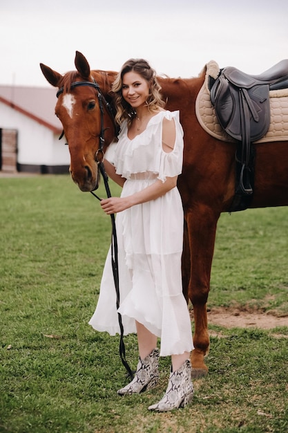 Belle fille dans une robe d'été blanche à côté d'un cheval sur un vieux ranch