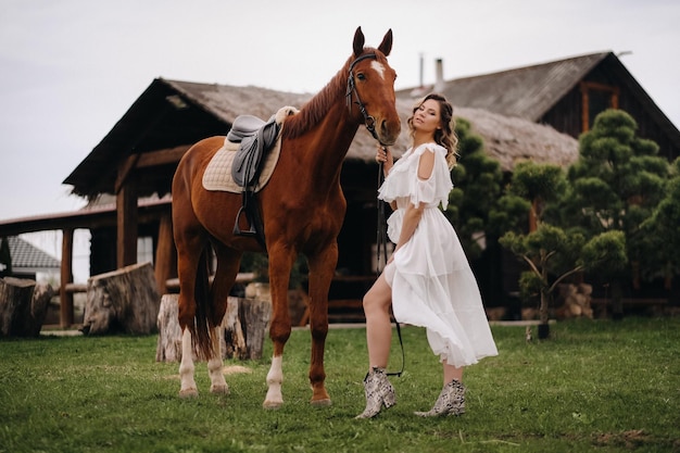 Belle fille dans une robe d'été blanche à côté d'un cheval sur un vieux ranch