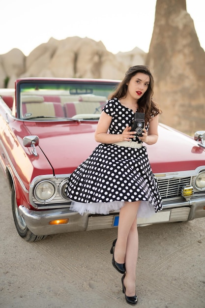 Belle fille dans un style rétro posant près d'une voiture cabriolet rouge vintage avec un vieil appareil photo argentique dans les mains