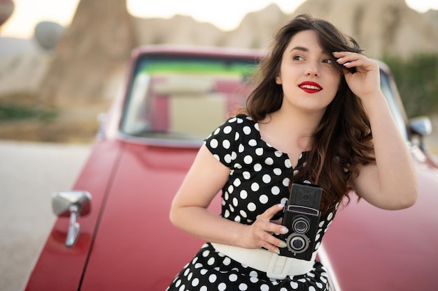 Belle fille dans un style rétro posant près d'une voiture cabriolet rouge vintage avec un vieil appareil photo argentique dans les mains