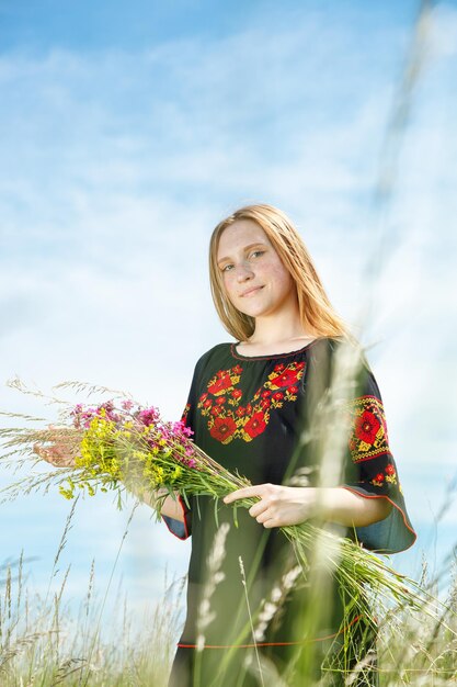 Belle fille dans un vyshyvanka noir avec des fleurs dans un champ avec vue sur le ciel