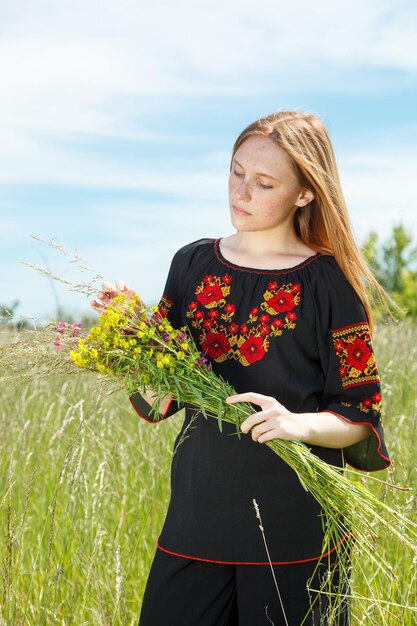 Belle fille dans un vyshyvanka noir avec des fleurs dans un champ avec vue sur le ciel