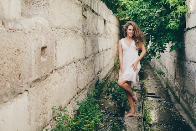 Belle fille dansante heureuse avec des cheveux naturels bouclés en robe blanche près des feuilles de l'arbre vert