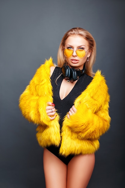 Belle fille dj à la mode avec des écouteurs en lunettes de soleil jaunes et manteau de fourrure jaune élégant sur fond gris