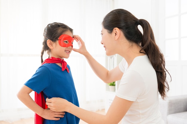 belle fille douce portant une cape rouge jouant comme super-héros et élégante jeune mère l'aidant à ajuster son masque dans le salon à la maison.