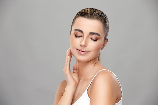 Belle fille avec du maquillage léger souriant avec ses yeux fermés et touchant sa joue sur un mur gris, concept de santé et de beauté