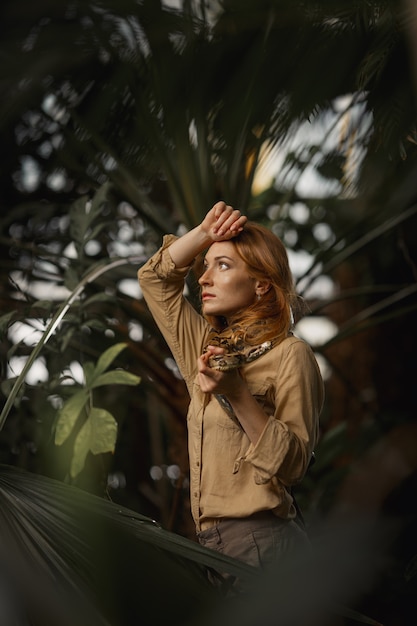 Une belle fille avec du maquillage naturel et des cheveux roux se tient dans la jungle parmi des plantes exotiques avec un serpent.