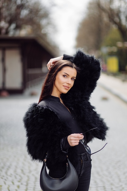 Belle fille élégante aux cheveux bruns en robe noire en plein air. Portrait de jeune femme élégante attrayante aux cheveux longs au printemps dans les rues de la ville.