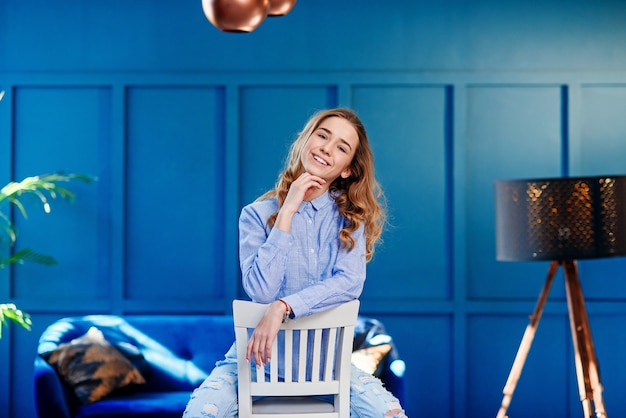 Belle fille élégante dans des vêtements décontractés est assis sur une chaise en bois blanche dans un intérieur moderne et élégant