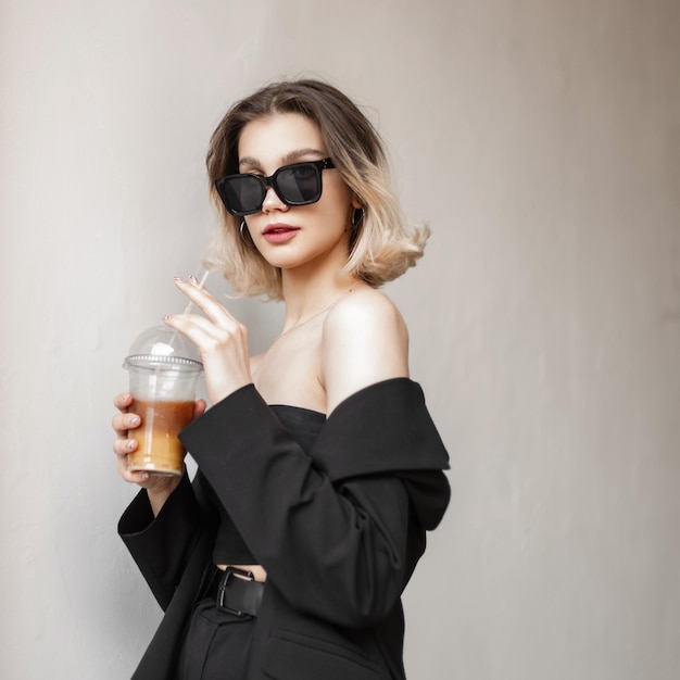 Belle fille élégante à la mode avec des lunettes de soleil glamour noires portant une tenue noire à la mode tient un café et marche dans la ville et se tient près du mur blanc