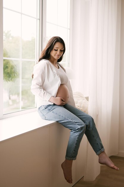 Belle fille enceinte avec des boucles dans une chemise blanche et un jean dans un studio lumineux avec un intérieur élégant le concept d'une grossesse et d'une famille heureuses