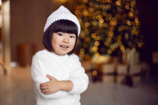 Belle fille enfant coréenne dans un pull et un chapeau tricotés blancs se tient à l'arbre de Noël le jour de Noël