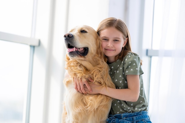 Belle fille enfant étreignant un chien golden retriever et souriant à l'intérieur enfant caressant un chien de race pure...