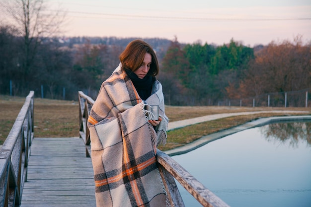 Une belle fille enveloppée dans un plaid se promène le long du pont au-dessus de l'étang avec une tasse de thé en métal par une froide journée d'automne