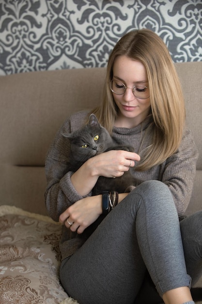 belle fille est assise à la maison sur le canapé et embrasse son chat gris aux yeux jaune vif