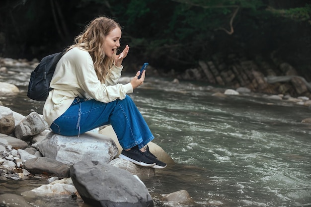 une belle fille est assise près d'une rivière de montagne, elle crie de colère dans le téléphone