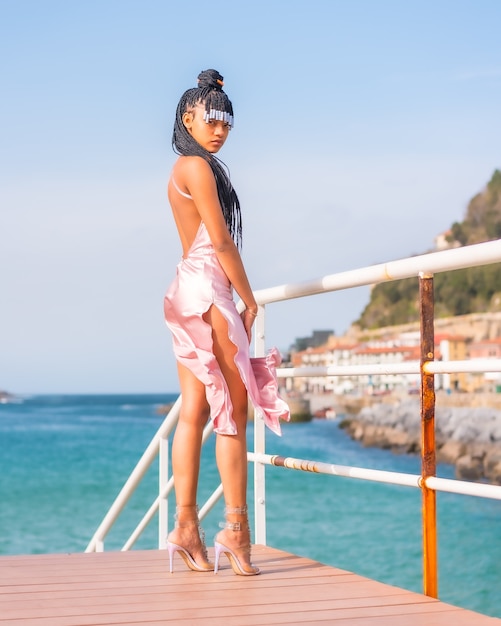 Belle fille ethnique dominicaine avec des tresses avec une belle robe rose. Mode profitant de l'été sur une passerelle en bois au bord de la mer