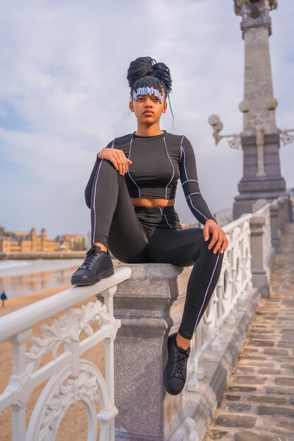 Belle fille ethnique dominicaine avec des tresses avec un costume complet. Mode, assis sur la balustrade de la plage en été