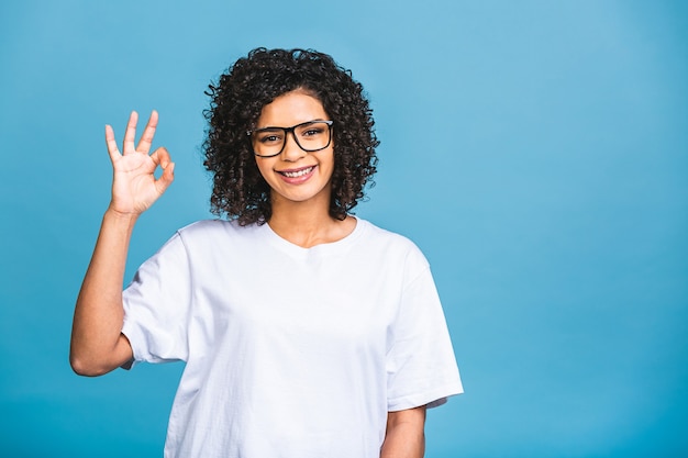 Belle fille étudiante afro-américaine avec une coiffure afro souriante isolée sur fond bleu. Ok signe.