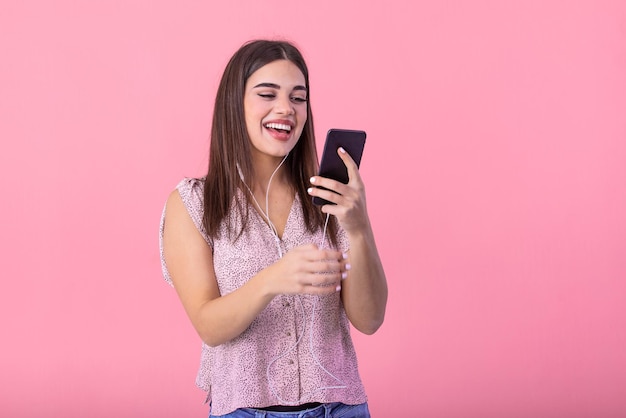 Une belle fille excitée tient un téléphone portable avec des écouteurs écoutant de la musique sur un écran vide isolé sur un fond de studio rose Emotions concept de style de vie Espace de copie