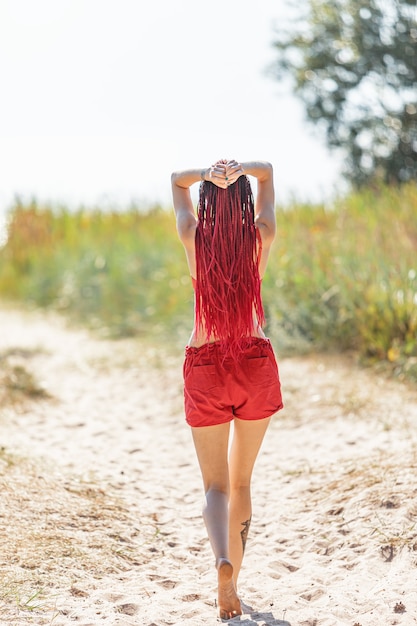 Belle fille à l'extérieur, profitant de la nature. Fille semi-nue avec des dreadlocks écarlates en maillot de bain rouge bronze sur la plage