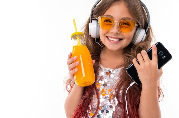 Belle fille avec gros écouteurs blancs tenant un smartphone noir et une bouteille de jus d'orange