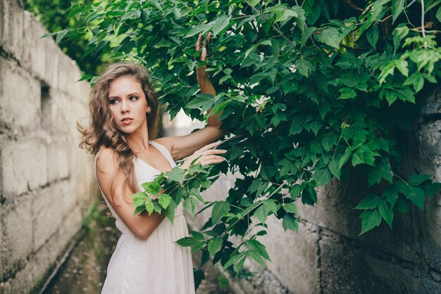 Belle fille heureuse aux cheveux naturels bouclés en robe blanche près des feuilles de l'arbre vert.