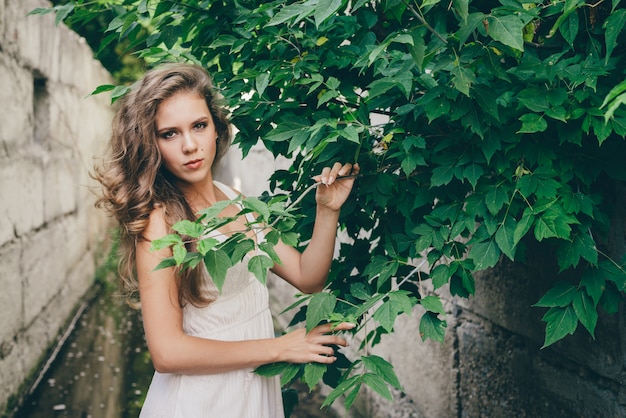 Belle fille heureuse aux cheveux naturels bouclés en robe blanche près des feuilles des arbres verts.