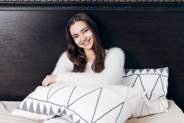 Belle fille heureuse en pyjama est assise dans son lit tôt le matin sous les yeux avec des taches blanches en souriant