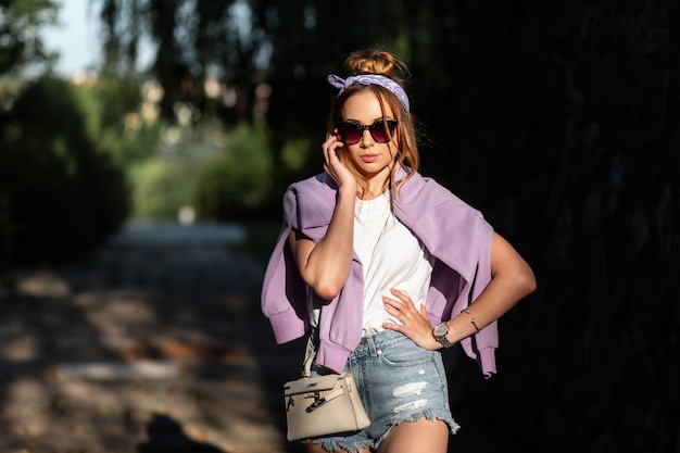 Belle fille hipster avec bandana et lunettes de soleil à la mode avec sweat-shirt et débardeur blanc en short en jean élégant avec sac à main marche dans la rue