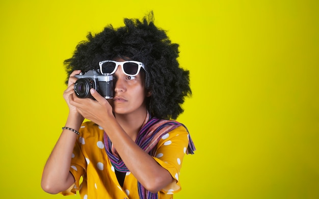 Belle fille indienne avec une coiffure frisée afro et des lunettes blanches parlant des photos avec appareil photo rétro sur mur jaune