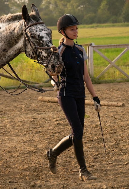 Belle fille jockey debout à côté de son cheval portant un uniforme spécial sur un fond de champ ciel et vert...