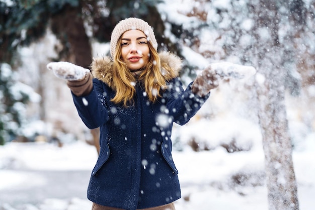 Belle fille jouant avec de la neige dans la forêt d'hiver Jeune fille souriante dans une veste bleue et un bonnet tricoté