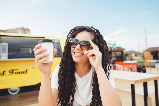 Une belle fille joyeuse avec des dreadlocks est assise sur une aire de restauration et boit du café café de restauration rapide dans la rue une femme boit du café dans un café automne