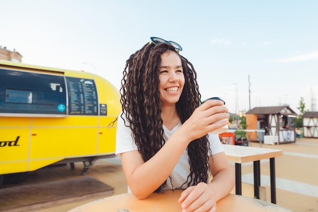 Une belle fille joyeuse avec des dreadlocks est assise sur une aire de restauration et boit du café café de restauration rapide dans la rue une femme boit du café dans un café automne