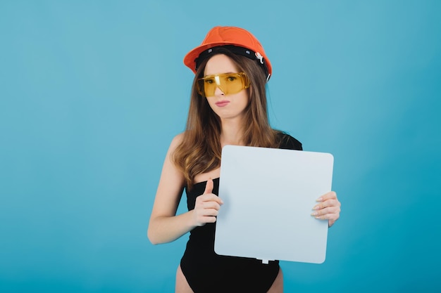 belle fille à lunettes jaunes et un casque de protection tient un tableau blanc dans sa main