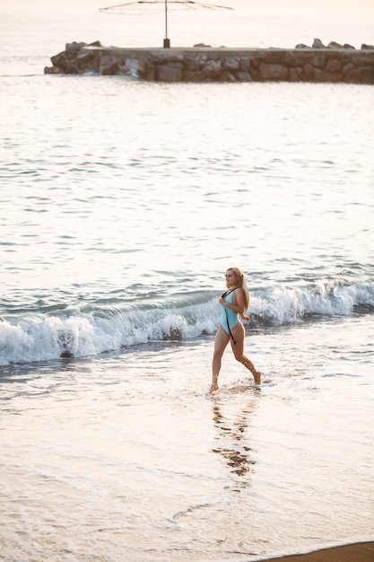 Belle fille en maillot de bain bleu et chapeau sur une plage de sable à la mer au soleil couchant