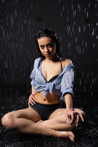Belle fille en maillot de bain noir et chemise est assise sous la pluie en profitant des gouttes d'eau Éclaboussure d'eau sur fond noir Aqua studio Posant et regardant la caméra