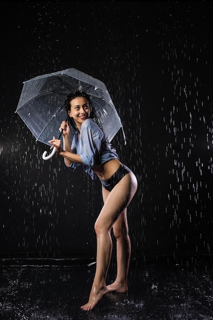 Belle fille en maillot de bain noir et chemise Se tient sous la pluie tient un parapluie transparent dans ses mains Regarde émotionnellement loin Éclaboussure d'eau sur fond noir Aqua studio