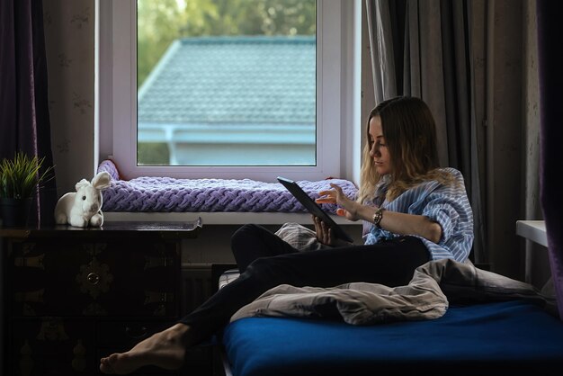 belle fille à la maison est assise sur le lit près de la fenêtre avec la lumière du jour et regarde dans la tablette