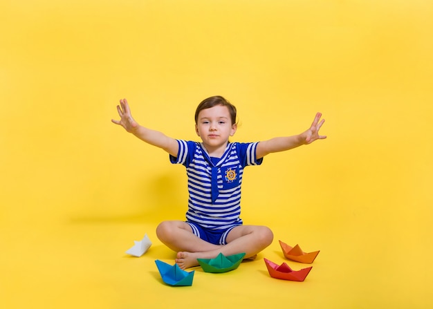 Une Belle Fille De Marin A étendu Ses Mains Sur Le Côté Et Est Assise Sur Un Espace Isolé Jaune. La Fille A Fait De L'origami En Papier. Bateaux En Papier Coloré.