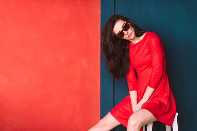 Belle fille de mode aux cheveux longs noirs, apparence espagnole en lunettes de soleil et robe élégante rouge posant sur un mur rouge bleu.