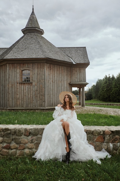 Belle fille modèle aux longues jambes dans une robe de mariée et un chapeau de paille posant près de l'église en bois dans la campagne.