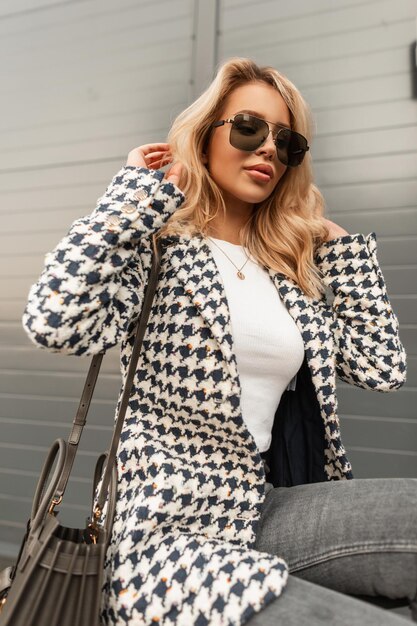 Belle fille modèle hipster avec des lunettes de soleil dans un manteau à la mode avec un t-shirt blanc et un sac dans la ville