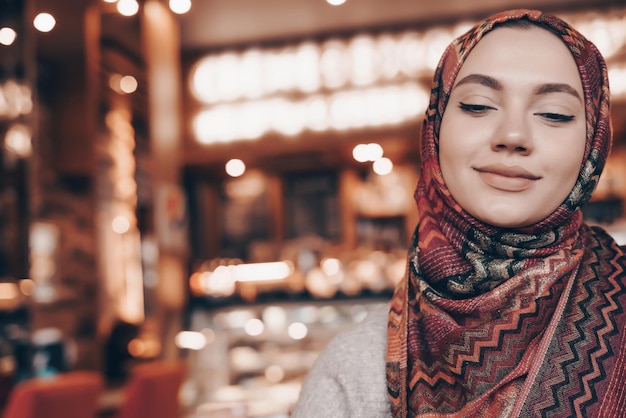 Une belle fille musulmane avec un foulard assis dans un café oriental confortable attendant sa nourriture et souriant