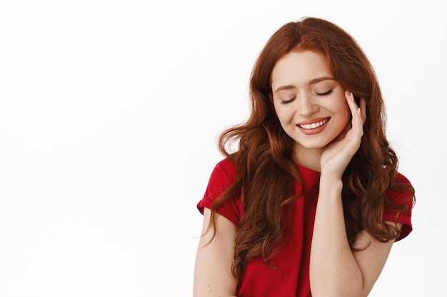 Belle fille naturelle aux cheveux roux et au sourire blanc, toucher la peau du visage fraîche et propre, rougissant et regardant vers le bas romantique, debout en t-shirt sur fond blanc.