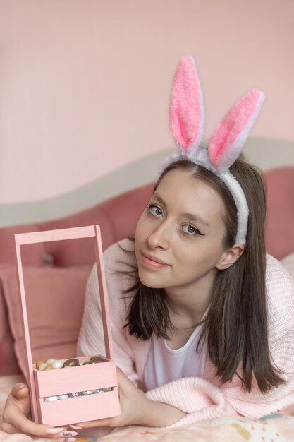 Belle fille avec des oreilles de lapin sur la tête se trouve sur le lit à la maison avec un panier d'oeufs de Pâques