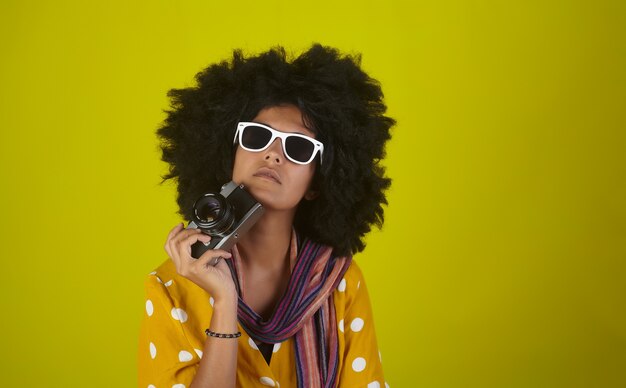 Belle fille de pensée avec une coiffure frisée afro tenant son appareil photo rétro sur un mur jaune