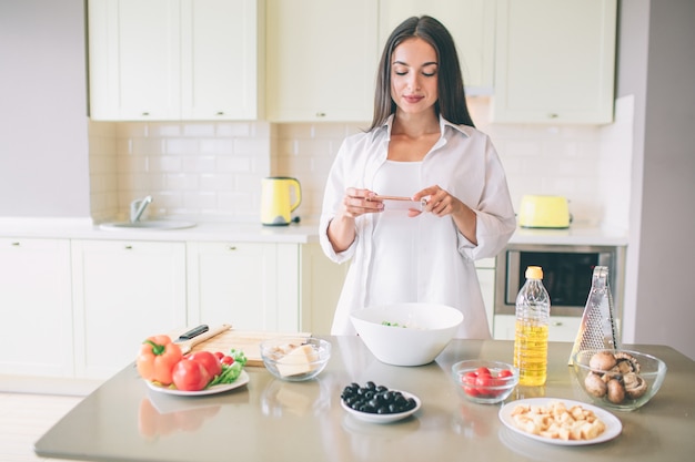 Belle fille ponce dans la cuisine et regarde le téléphone qu'elle tient dans les mains. La fille a arrêté de cuisiner. Elle a une pause.