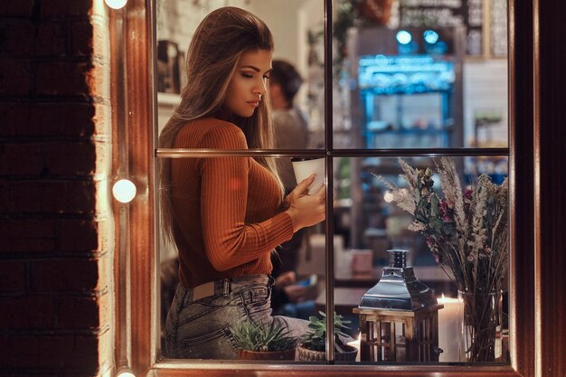 Une belle fille portant un pull tenant un café assis sur un rebord de fenêtre à l'intérieur du café